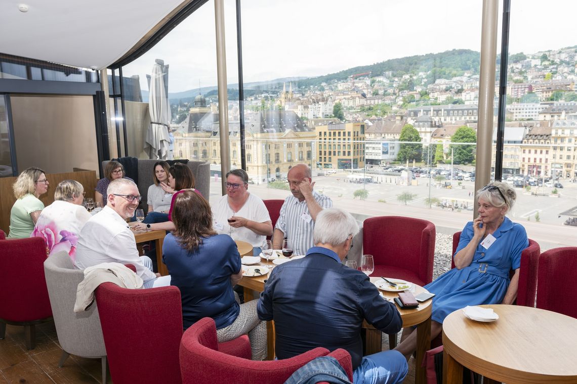 Mehrere Leute sitzen an Tischen. Durch ein grosses Fenster ist die Stadt Neuenburg sichtbar.