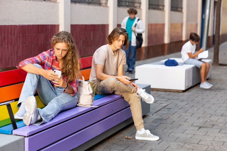 Eine Teenagerin und ein Teenager sitzen zusammen auf einer Bank vor einer Schule.