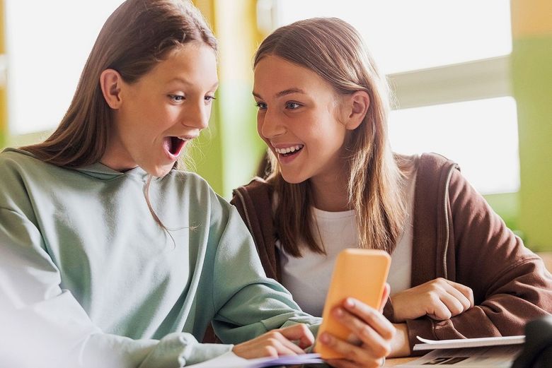 Eine Jugendliche betrachtet mit offenem Mund etwas auf einem Smartphone. Eine weitere Jugendliche beobachtet sie grinsend.