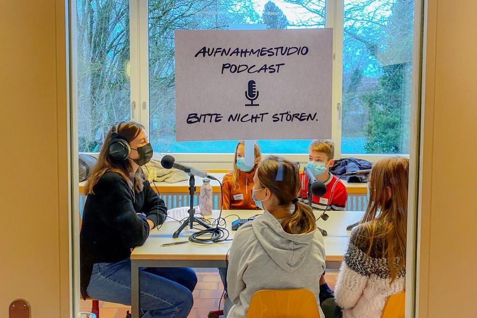 Schild an Glastüre bittet um Ruhe wegen Podcast-Aufnahmen – im Zimmer sitzen eine Moderatorin und Kinder mit Mikrofonen am Tisch.