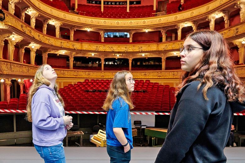 Drei Schulkinder stehen auf der Opernbühne. Im Hintergrund sind Sitzreihen sichtbar.