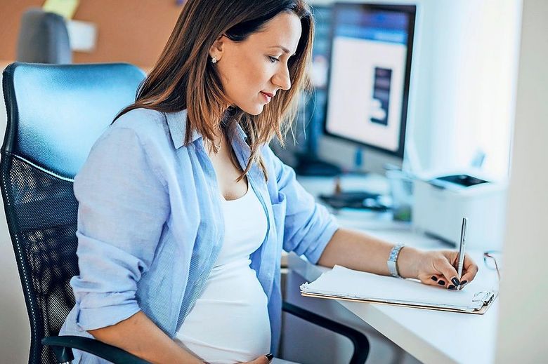 Am Bürotisch sitzt eine schwangere Frau, die sich vor dem Computer Notizen macht.