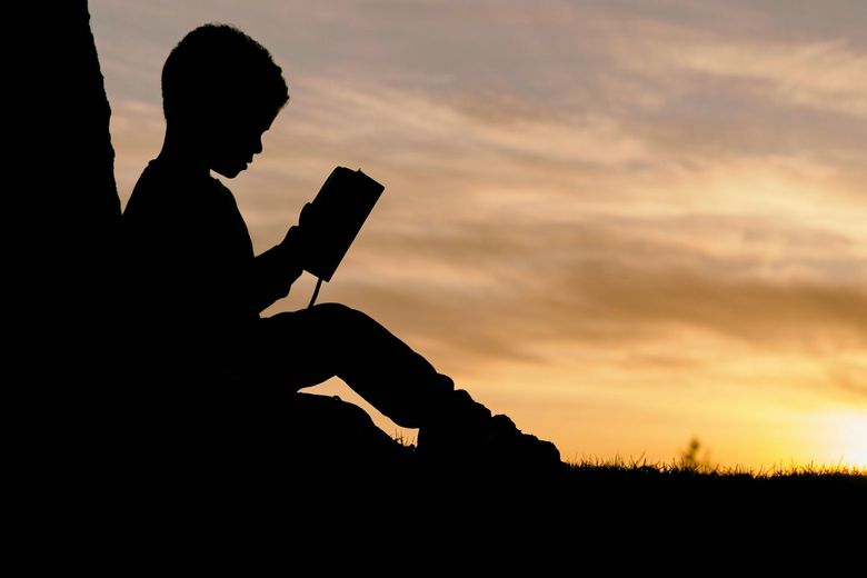 Die Silhouette eines Kindes, das sitzt und ein Buch liest. Im Hintergrund steht die Sonne tief am Horizont.