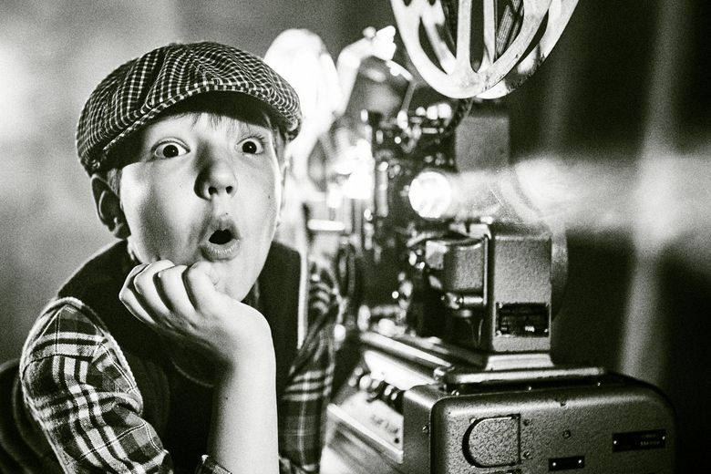 Retro-Symbolbild: Ein Junge mit Mütze sitzt staunend neben einem alten Filmprojektor. 