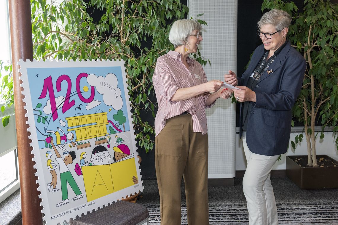 Antoinette Killias übergibt Rita Marty einen Umschlag. Neben Killias befindet sich das Bild einer Briefmarke.