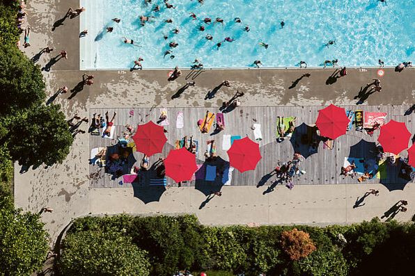 Schwimmende in einem Freibad. Auf der Wiese sind knallrote Sonnenschirme aufgespannt.