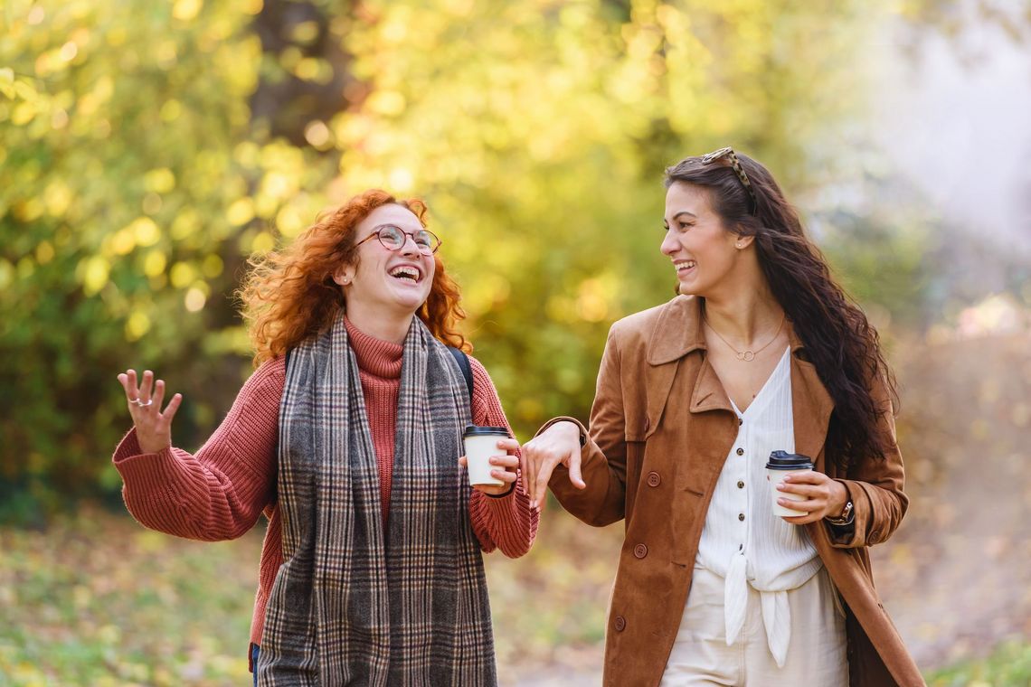 Zwei fröhliche junge Frauen spazieren in einem Park und halten je einen Kaffeebecher in der Hand.