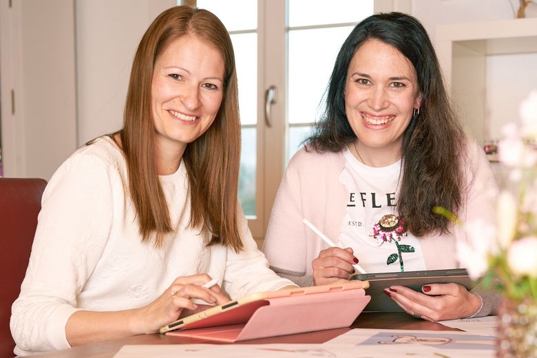 Zwei Frauen sitzen lächelnd an einem Tisch.