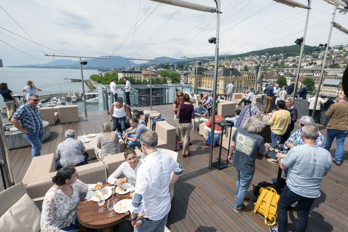 Mehrere Leute befinden sich auf einer Dachterrasse, sprechen miteinander und trinken. Im Hintergrund sind das Ufer eines Sees sowie eine Stadt zu sehen.