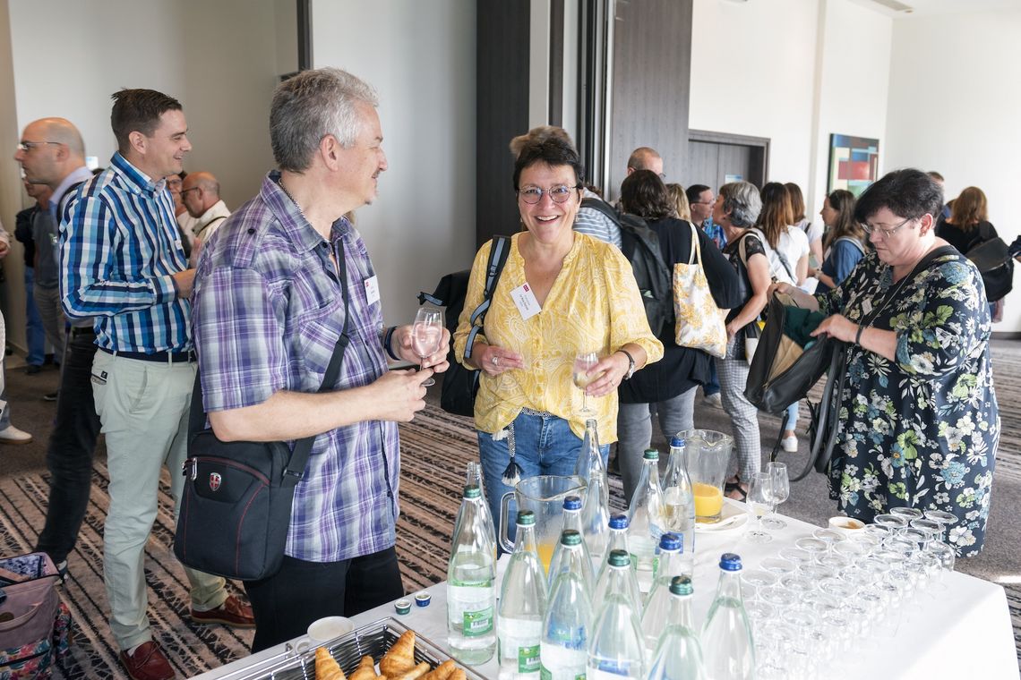 Eine Frau steht vor einem Tisch mit Getränken und lacht in die Kamera. Um sie herum befinden sich mehrere Leute. Foto: Marc Renaud