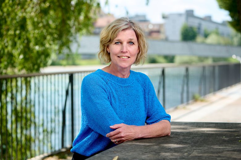 Blonde Frau sitzt an einem Fluss und blickt in die Kamera.