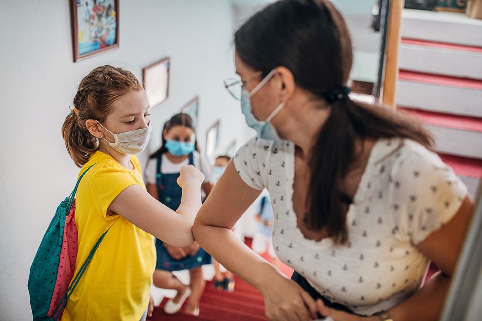 Lehrerin grüsst Schülerin mit dem Ellbogen. Beide tragen Masken.