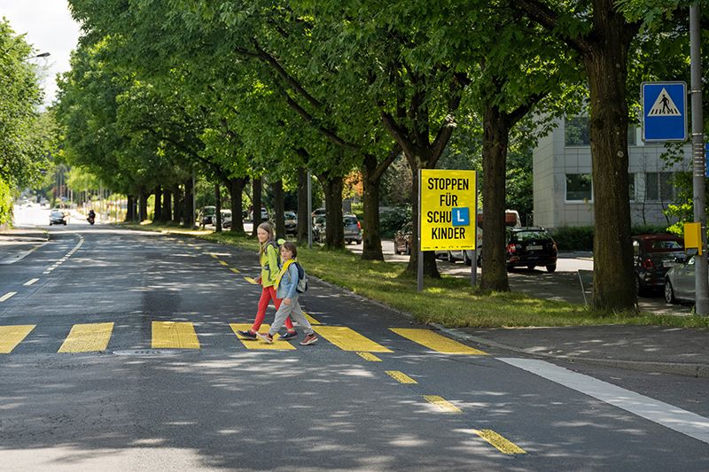 Schulkinder überqueren einen Zebrastreifen, im Hintergrund ist das Plakat der Kampagne zu sehen. 