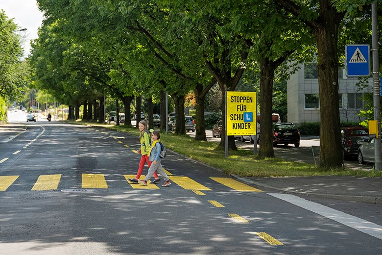 Schulkinder überqueren einen Zebrastreifen, im Hintergrund ist das Plakat der Kampagne zu sehen. 
