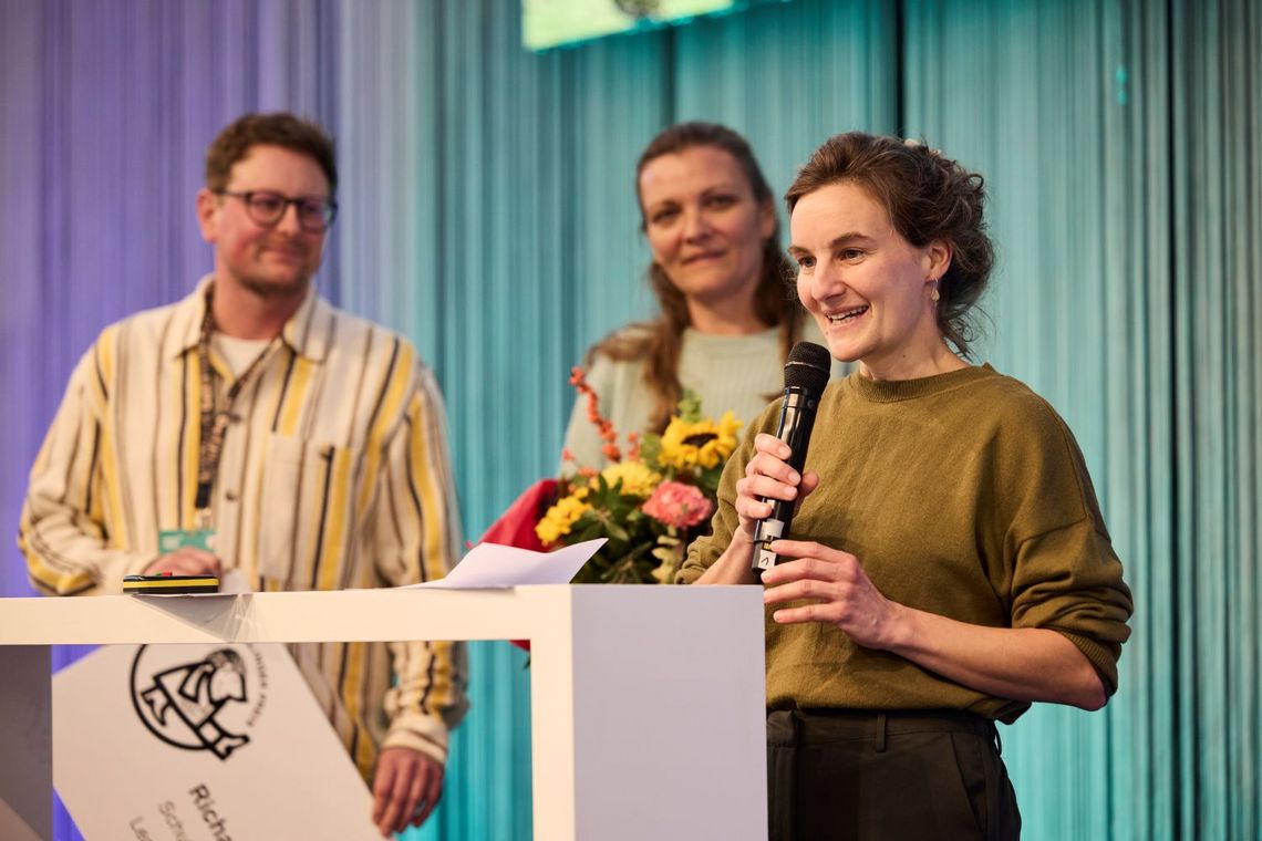 eine Frau spricht in ein Mikrofon, zwei weitere Personen hören ihr zu.