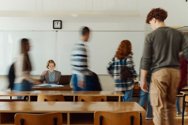 Schülerinnen und Schüler schwirren um eine Lehrerin, die am Tisch vor der Wandtafel sitzt. 